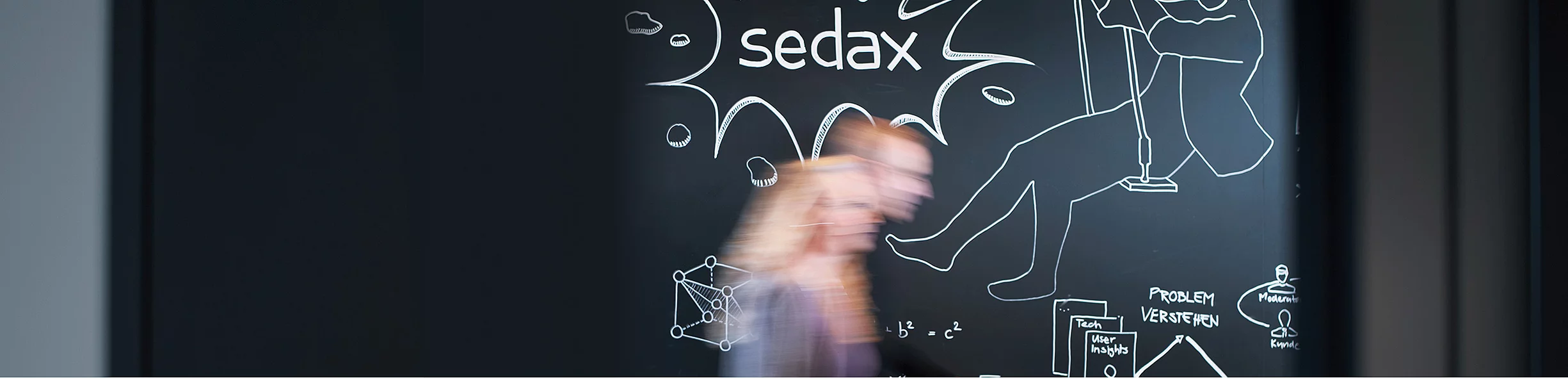 Person geht unscharf an einer Tafel mit Kreide-Skizzen vorbei, darauf eine Sprechblase mit dem Schriftzug "sedax" sowie Notizen und Zeichnungen wie "Problem verstehen"