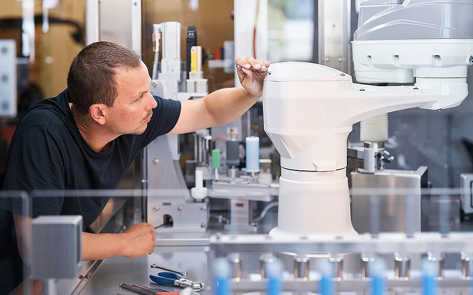 Mitarbeiter justiert einen Roboterarm in einer automatisierten Produktionszelle, im Hintergrund Fläschchen auf einem Förderband