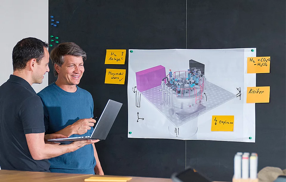 Zwei Mitarbeiter besprechen ein 3D-Konstruktionsmodell an einer Wand mit Notizzetteln