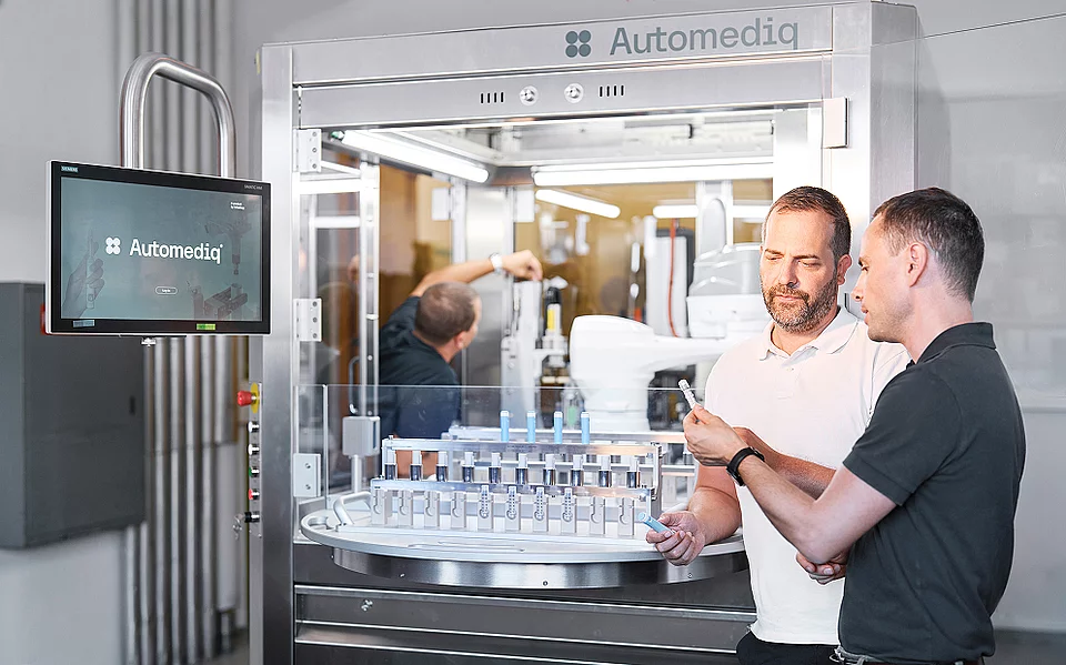 Zwei Mitarbeiter besprechen ein Bauteilraster mit Fläschchen vor der Automediq-Produktionsanlage, im Hintergrund ein Roboterarm