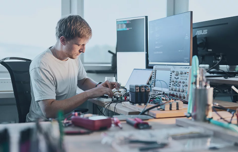 Mitarbeiter arbeitet an einer elektronischen Schaltung am Labortisch, im Hintergrund Bildschirme mit Messdaten und ein Oszilloskop
